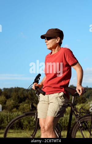 Eine Frau mittleren Alters in einem roten T-Shirt und Mütze ruht am Fahrrad und genießt das schöne sonnige Wetter. Stockfoto