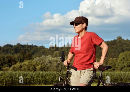 Eine Frau mittleren Alters in einem roten T-Shirt und Mütze ruht am Fahrrad und genießt das schöne sonnige Wetter. Stockfoto