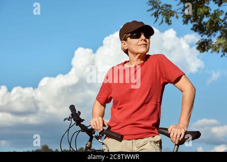 Eine Frau mittleren Alters in einem roten T-Shirt und Mütze ruht am Fahrrad und genießt das schöne sonnige Wetter. Stockfoto