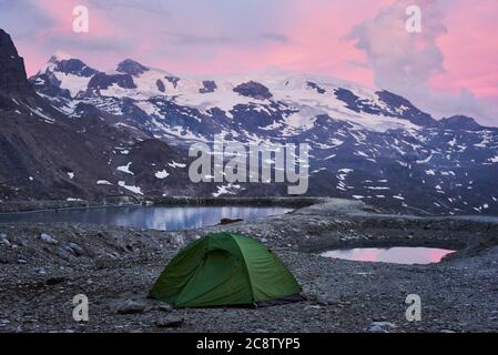 Grünes Touristenzelt in leeren felsigen Bergen in der Nähe von kleinen Seen mit frischem klarem Wasser. Alpine Berge leuchten durch rosa Sonnenuntergang Licht. Beliebte Touristenattraktion. Konzept der Natur Schönheit, Camping. Stockfoto