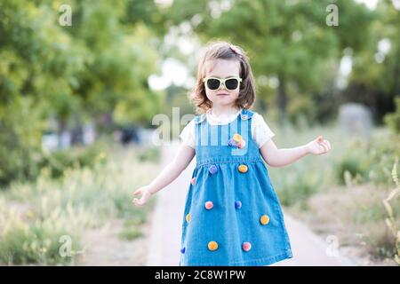 Stilvolle Baby Mädchen 2-3 Jahre alt trägt Denim-Kleid und Sonnenbrillen im Freien im Park. Blick auf die Kamera. Sommersaison.