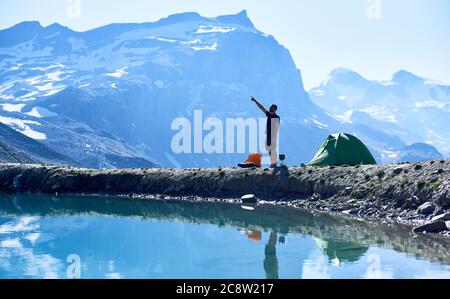 Seitenansicht des Reisenden in kurzen Hosen, die auf Berge zeigen, während sie am wunderschönen blauen See stehen. Tourist Zelt und junger Mann in kristallklarem Wasser reflektiert. Konzept von Reisen, Wandern und Camping. Stockfoto