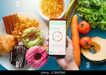 Kalorienzählen und Lebensmittelkontrolle Konzept. Frau mit Kalorienzähler Anwendung auf ihrem Smartphone mit frischem Gemüse, Dessert und Donuts Stockfoto