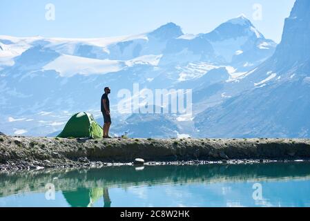 Seitenansicht des Mannes, der Landschaft genießt, stehend nahe Zelt in leeren felsigen alpinen Bergen nahe See mit frischem klarem Wasser. Beliebte Touristenattraktion in sonnigen warmen Sommertag. Konzept der Schönheit der Natur. Stockfoto