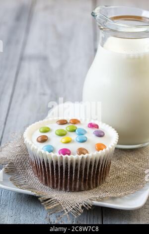 Schokoladen-Cupcake mit weißer Verglasung und bunten Smarties auf einem Teller und Glas Milch, Holzhintergrund Stockfoto