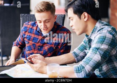 Vietnamesischer junger Mann zeigt neue mobile Anwendung auf seinem Smartphone an Freund, wenn sie sitzen im Café tble Stockfoto