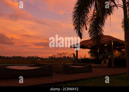 Schöner orange feuriger Sonnenuntergang in den Tropen. Straßendach von Heu, neben dem Pool und mit Blick auf das Reisfeld Stockfoto