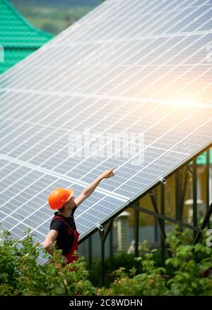 Mann Arbeiter in einem orangen Helm zeigt auf eine hohe Solarstation auf einem Grundstück mit viel Grün. Alternative billige Sonnenenergie-Produktion und profitables finanzielles Investitionskonzept Stockfoto