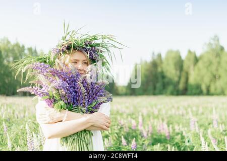 Ein glückliches Mädchen in einem Blumenkranz umarmt einen Strauß wilder Blumen und lächelt, atmet den Duft von Blumen, ein Bouquet von Lupinen. Glücklich fröhlich Stockfoto