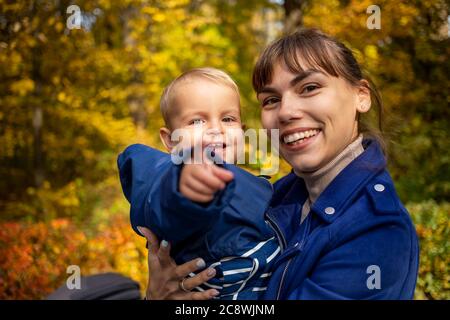 Junge schöne Mutter und Kleinkind auf ihren Händen schauen auf die Kamera und lachen glücklich im Herbstpark Stockfoto