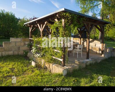 Brown tiber Holzpavillon oder Pergola mit Kletterpflanzen und Blumen, Tisch und Stühle und umliegenden Sandsteinwand, grünes Gras blauen Himmel in Stockfoto