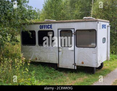 Kleines weißes Wohnwagenhaus mit blauem Schild, verlassene Wohnanlage in ländlicher Landschaft in der Nähe von Fußweg, grünem Gras und Bäumen Stockfoto