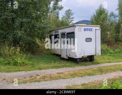 Kleines weißes Wohnwagenhaus mit blauem Schild, verlassene Wohnanlage in ländlicher Landschaft in der Nähe von Fußweg, grünem Gras und Bäumen Stockfoto