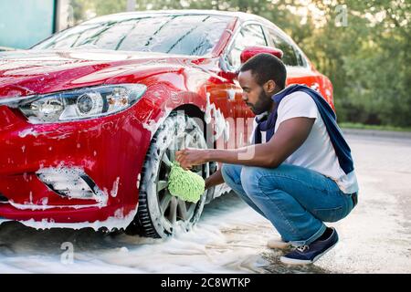 Luxus rotes Auto in Seifenschaum im Freien bei Autowaschservice.  Seitenansicht des hübschen jungen afrikanischen Mann mit grünen Mikrofaser  Auto Waschhandschuh für die Reinigung Stockfotografie - Alamy