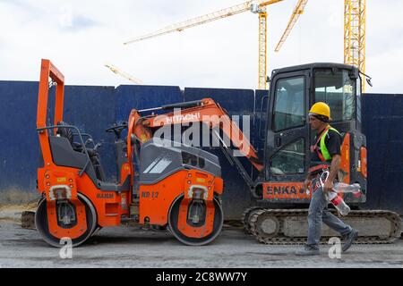 Bauarbeiter, der durch einen roten Bagger und Walzenverdichter geht, der in der Nähe einer Baustelle geparkt ist, Krane im Hintergrund. Stockfoto