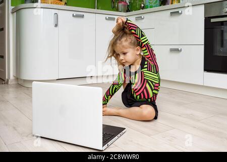 Kleines Mädchen in einem Turnanzug ist in rhythmische Gymnastik online zu Hause beschäftigt und schaut auf einen Laptop Stockfoto