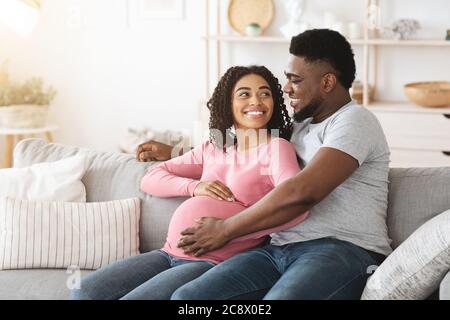 Schöne glücklich erwartete afrikanische Eltern sitzen auf dem Sofa zu Hause Stockfoto