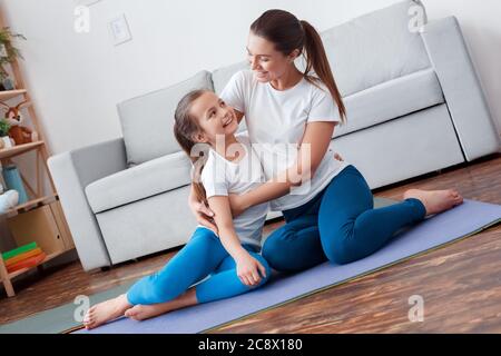 Gesunder Lebensstil. Mutter und Tochter in Sportkleidung sitzen auf Yoga-Matte umarmt lächelnd glücklich Stockfoto