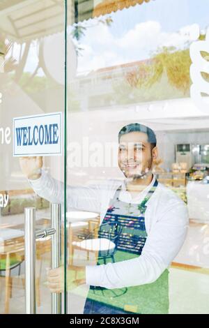Lächelnder kleiner Bäckerbesitzer, der morgens ein Begrüßungsschild an der Glastür aufstockt, wenn er das Café öffnet Stockfoto