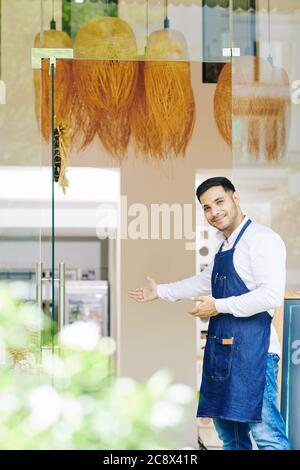 Positiver Cafe Kellner in Denim Schürze, der an Eingangstüren steht und Gäste einlädt Stockfoto