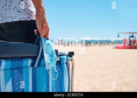 Nahaufnahme eines kaukasischen Mannes, der kurze Hosen trägt, eine OP-Maske und einen gefalteten Strandstuhl auf dem Weg zum Strand trägt, mit dem Meer im Hintergrund Stockfoto