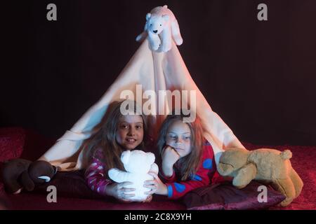 Kinder in Schlafanzug mit Decke bedeckt spielen mit Teddybären. Kinder haben Pyjama-Party mit Spielzeug auf schwarzem Hintergrund. Mädchen mit lächelnden Gesichtern unter der Decke Zelt. Kindheit und Glück Konzept Stockfoto