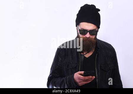 Street Life Style Konzept. Typ mit Bart in schwarzer Lederjacke und Hut. Rock Fan oder Gangster in Sonnenbrille hält Handy. Mann mit geschäftigen Gesicht und brutalen Stil isoliert auf weißem Hintergrund Stockfoto