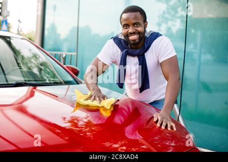 Porträt eines lächelnden jungen dunkelhäutigen Mannes, der seine rote Autohaube mit gelbem Mikrofasertuch im Freien an der Autowaschstation putzt Stockfoto