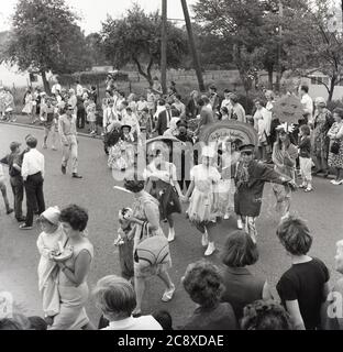 1967, historisch, bei einem englischen Dorffest in Bierton, Buckinghamshire, England, beobachten eine Menge einheimischer Leute, die am Straßenrand stehen, wie Kinder in Kostümen gekleidet sind, mit einem jungen Mädchen, das als „britisches Wetter“ verkleidet ist, und anderen als „Mitternacht“, „Mittag“ und „Heilig“. Stockfoto