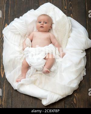 Kleinkind mit weißer Decke bedeckt. Kindheit und Unschuld Konzept. Baby liegt auf weißen weichen Bettdecke als Wolke oder Nest entworfen. Neugeborenes Kleinkind mit blauen Augen und neugierigem Gesicht auf Holzhintergrund Stockfoto