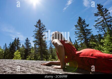 Junge langhaarige Frau in einem ärmellosen leuchtend roten Stadtkleid auf einem Felsen in der Mitte der kanadischen Wälder liegen. Verbindung mit der Natur. Sonneneruptionen. Stockfoto