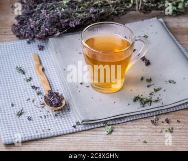 Warmer Oregano-Tee in einer Glasschale steht auf Leinen beige Servietten auf dem Hintergrund von trockenem Oregano. Ein kleiner Holzlöffel mit trockenen Oregano Blüten liegt nea Stockfoto