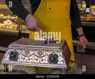 In einem Café macht ein Barista in einer gelben Schürze türkischen Kaffee auf Sand in einem türken. Stockfoto