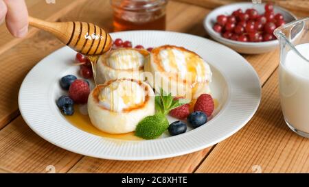 Gießen von Honig auf Quark fritters. Russische ukrainische Küche Essen Syrniki, süße Käsesorten mit saurer Sahne, Beeren und Honig serviert Stockfoto