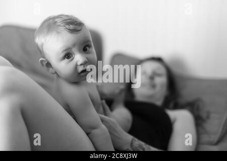 Mutter und Baby Sohn freudige Intimität. Baby auf dem Bauch seiner Mutter sitzend und mit einem lustigen Ausdruck auf die Kamera schauend. Schwarz-Weiß-Porträt. Stockfoto