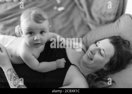 Mutter und Baby Sohn freudige Intimität. Baby liegt auf der Brust seiner Mutter und schaut mit einem lustigen, überraschenden Ausdruck auf die Kamera. Schwarz-Weiß-Bild Stockfoto