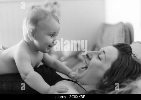 Mutter und Baby Sohn freudige Intimität. Baby liegt auf der Brust seiner Mutter und schaut auf seine Mutter. Beide lächeln und lachen. Schwarz-Weiß-Porträt. Stockfoto