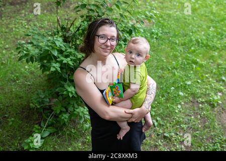 Porträt einer Mutter und ihres Babys, im Freien im Wald. Mama hält ihren Sohn in den Armen, beide posieren für Kamera, Baby mit lustigen Ausdruck. Stockfoto