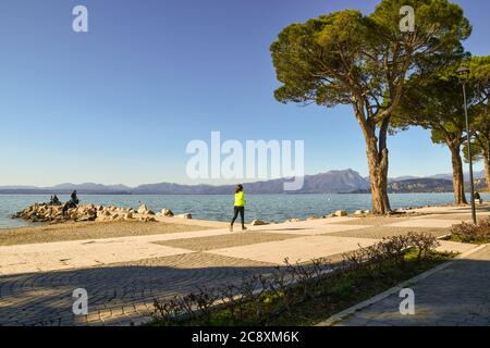 Landschaftlich schöner Blick auf den Gardasee mit einer Frau, die auf der Seepromenade läuft und Menschen auf den Felsen an einem sonnigen Wintertag, Lazise, Verona, Venetien, Italien Stockfoto