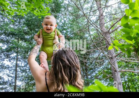 Porträt einer Mutter und ihres Babys, im Freien im Wald. Mama hält ihren Sohn in den Armen hoch, während Baby lacht und anbetungsvoll auf die Kamera schaut. Stockfoto