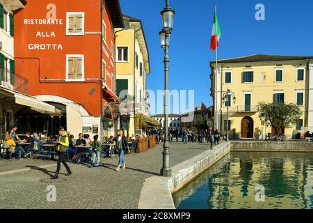 Blick auf die Altstadt am Ufer des Gardasees mit Menschen zu Fuß und sitzen in Outdoor-Restaurants und das Rathaus, Lazise, Venetien, Italien Stockfoto