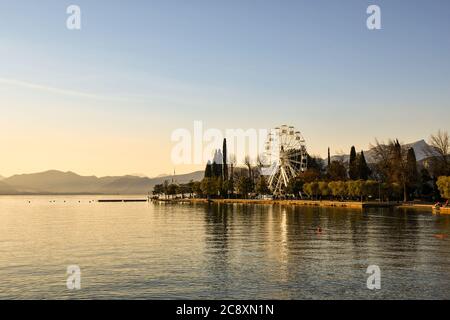 Panoramablick auf den Gardasee bei Sonnenuntergang mit einem Panoramarad auf der Seepromenade und der Küste im Hintergrund, Bardolino, Venetien, Italien Stockfoto