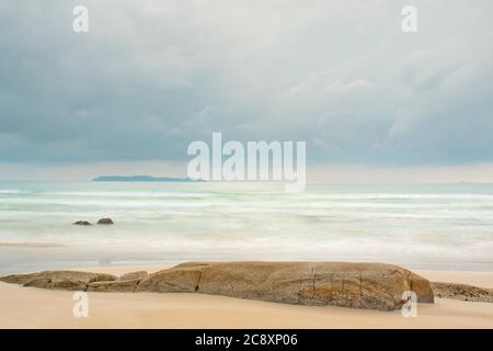 Ruhige Szene eines Strandes am Meer mit einer Insel im Hintergrund und Felsen im Vordergrund - pastellfarben durch lange Belichtungszeit Stockfoto