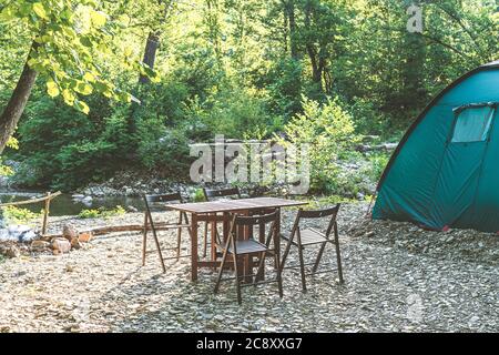 Camping am Strand des Bergflusses. Blaues Zelt, Lagertisch und Stühle im Wald. Sommerreisethema. Ruhestätte in der Natur. Stockfoto