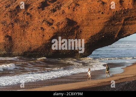 Ein verliebten Paar, das bei Sonnenuntergang unter dem riesigen Bogen am Strand von Legzira spazieren geht. Berühmtes und beliebtes Wahrzeichen in Marokko. Stockfoto