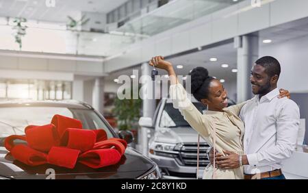 afrikanerin ist glücklich, nachdem sie ein Auto von ihrem Mann bekommen hat, sie schaut ihn mit Liebe und Dankbarkeit an. Neues Auto mit rotem Bogen im Hintergrund Stockfoto