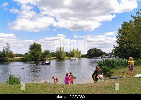 Die Themse in Chertsey mit Kindern, die an einem Sommertag auf dem Damm spielen, während im Hintergrund Flussboote kreuzen, Surrey England Stockfoto