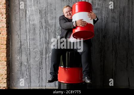 Lächelnder fett kaukasischen Geschäftsmann ist nicht bereit, jemandem seinen Gewinn auf die Ölproduktion geben, umarmt Mann roten Ölfass Stockfoto