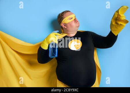 Portrait von lustigen fröhlichen fetten Mann mit Waschmittel in den Händen, ist der Mann bereit zu reinigen, trägt gelbe Kostüm, Handschuhe und Maske von Superhelden. Isoliert blau Stockfoto