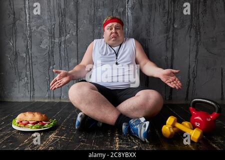 Fettleibiger Kaukasischer Mann kann nicht entscheiden, ob zu trainieren oder zu essen, sitzt männlich auf dem Boden und wählen Sie ungesunde Lebensmittel zu essen oder Sport-Übungen Stockfoto